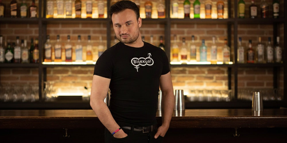 A man wearing a black t-shirt with the word bisexual on the chest, standing in an uplit bar