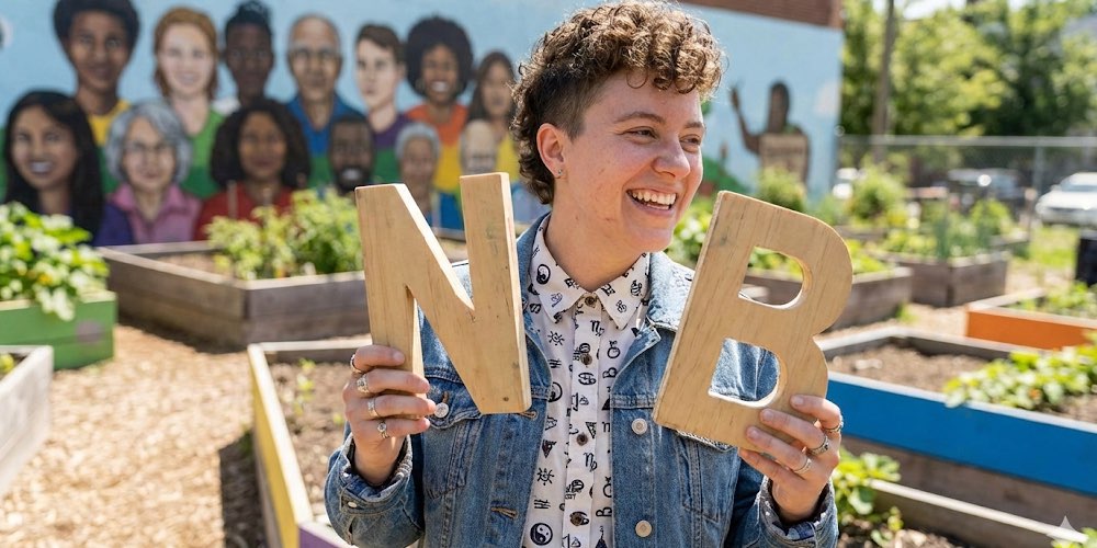 A non-binary person in an urban allotment, holding wooden letters N and B