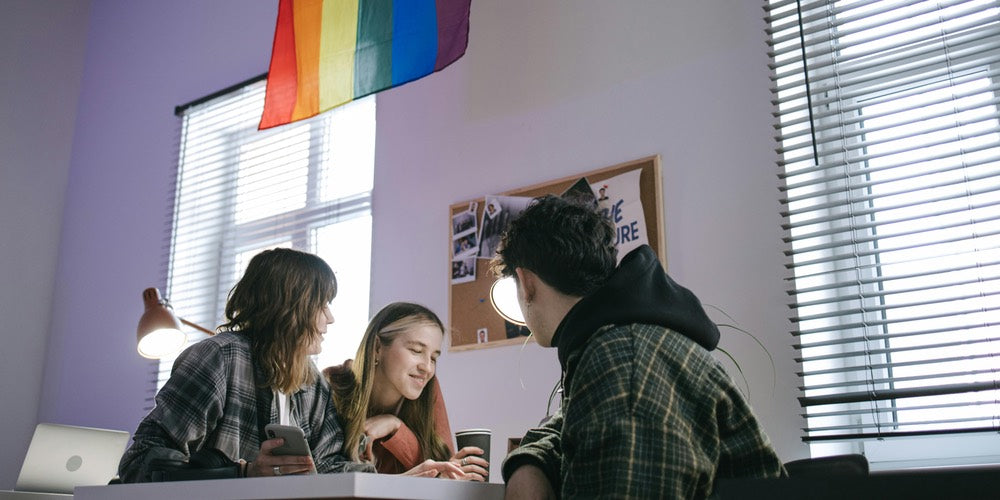 Three LGBTQ+ people working in an office, with a rainbow pride flag on the wall