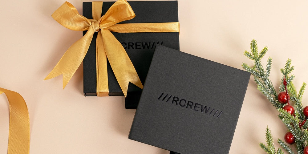 Two black gift boxes with RCREW branding, one with a yellow ribbon. Surrounded by pine twigs and berries