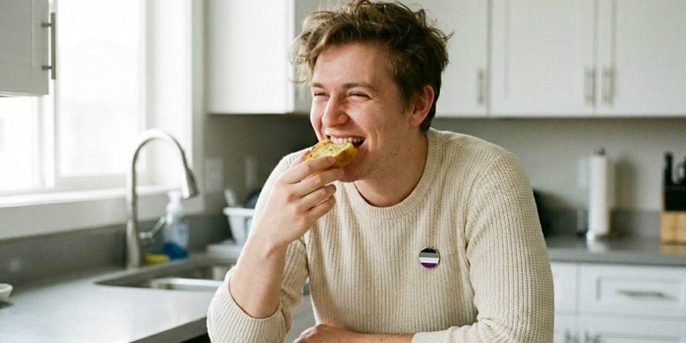 A young man in a kitchen wearing an asexual pride flag pin on his jumper, eating a piece of garlic bread